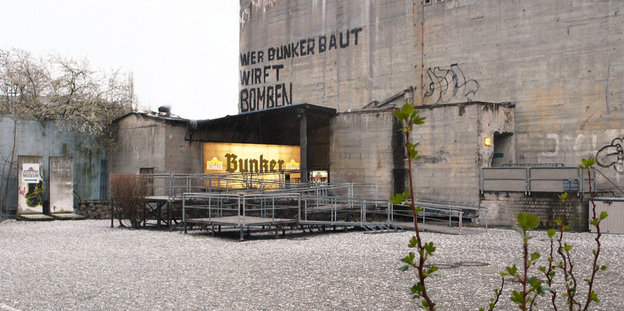 Ein Hof und ein Bunker. Auf dem Bunker steht ein Graffito: "Wer Bunker baut, wirft Bomben."