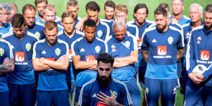 viele Spieler in blauen Trikots stehen auf dem Rasen, vor ihnen einer mit Bart, es ist Jimmy Durmaz