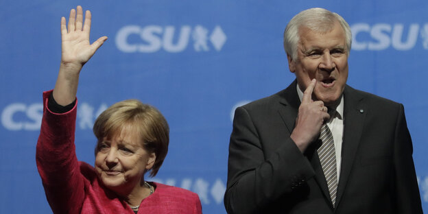 Angela Merkel winkt, während Horst Seehofer sich ins Gesicht fasst