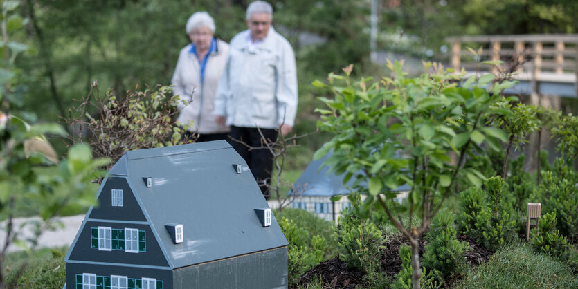 Im Vordergrund steht ein Miniaturhaus, im Hintergrund läuft ein älteres Paar