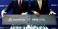 Zwei Personen in Anzügen halten Schilder mit den Logos der Unternehmen Thyssenkrupp und Tata Steel