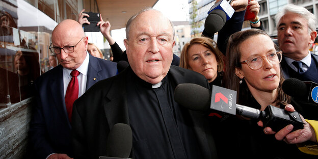 Der verurteilte Erzbischof Philip Wilson befindet sich in der Mitte des Bildes. Er verlässt das Amtsgericht in Newscastle. Um ihn herum stehen Reporter, sie halten Mikrofone in seine Richtung.