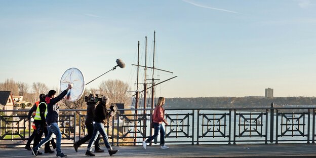 Frau in roter Jacke geht über eine Brücke, gefolgt von einem Kamerateam