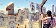 Eine Frau hängt ein Vermisstenplakat an einem Zaun auf, hinter dem ein Stein mit der Aufschrift „Villa Baviera“ steht