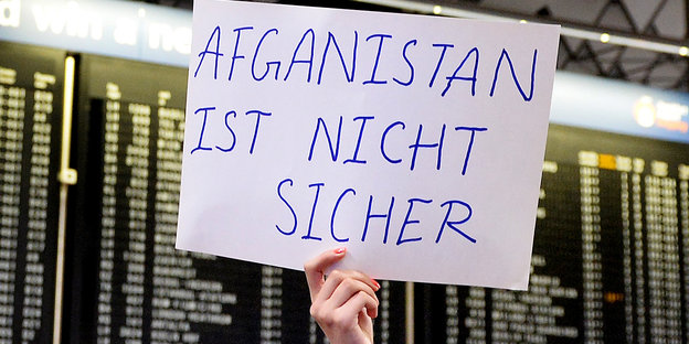 Vor einer Anzeigetafel am Flughafen hält eine Person ein Schild „Afghanistan ist nicht sicher“