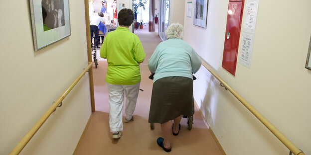 Eine Pflegerin und eine alte Frau mit Rollator gehen über einen Flur