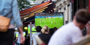 Menschen stehen zur Fußball WM vor einem Bildschirm und gucken dem Spiel zu