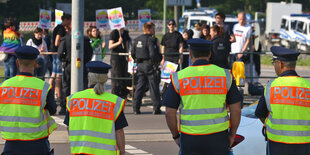 Polizist*innen stehen mit neongelben Warnwesten bekleidet an einer Absperrung. Dahinter sieht man Demonstranten