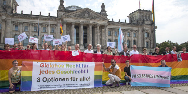 Eine Menschenmenge steht vor dem Bundestag. Sie halten eine Regenbogenflagge und Banner hoch. Die Menschen lächeln in die Kamera.