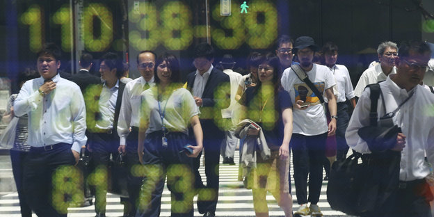 Menschen laufen über eine Straße in Tokio, es spiegeln sich Zahlen in einer Scheibe