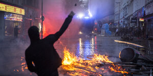 Eine Person wirft einen Stein auf einen Polizeiwagen hinter einer brennende Barrikade