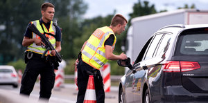 Zwei Polizisten stehen neben einem Auto. Sie tragen Warnwesten, einer beugt sich zum Fenster der Fahrerseite hinunter. der andere hält eine Waffe in den Händen