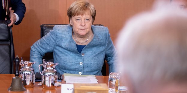 Angela Merkel schaut mit strenger Miene auf einen weißhaarigen Mann (Horst Serehofer), der ihr gegenüber sitzt und nur von hinten zu sehen ist.
