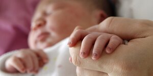 Ein neugeborenes Baby hält die Hand eines Erwachsenes. Das Gesicht des Babys ist unscharf im Hintergrund zu erkennen.