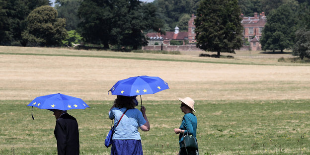 Drei Menschen stehen neben einem Feld, im Hintergrund ist Chequeurs, der offizielle Landsitz der britischen Premierministerin, zu sehen. Zwei der drei Personen halten blaue Sonnenschirme mit gelben Sternen in der Hand - das EU-Logo