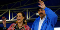 Rosario Murillo, in buntem Kleid und mit vielen Ringen an der Hand, und Daniel Ortega in blauer Strickjacke winken von einer Bühne.