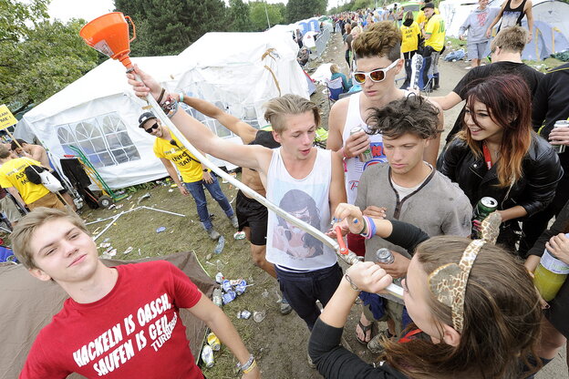 Auf einem Festivalgelände trinkt ein Mädchen aus einem Trichter mit Gartenschlauch, andere Jugendliche schauen zu und amüsieren sich.
