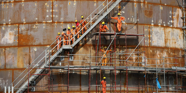 Bauarbeiter in orangefarbenen Anzügem stehen auf einer Leiter, die Außen an dem rostigen Tank einer Ölraffinerie hinaufführt