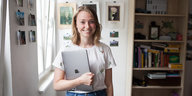 Eine lächelnde junge Frau mit Laptop unter Arm steht neben einem Bücherregal