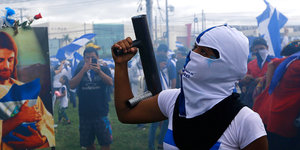 Im Vordergrund eine maskierte Demonstrantin, die einen selbstgebauten Granatwerfer in der Hand hält. Im Hintergrund Menschen mit blau-weißen nicaraguanischen Fahnen.