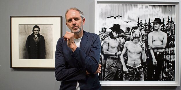 EIn Mann steht vor einer grauen Wand, an der schwarz-weiß-Fotos hängen
