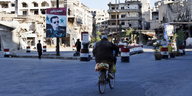 Im syrischen Homs fährt ein Mann auf dem Fahrrad auf einen Kontrollposten zu. Im Hintergrund ausgebombte Häuser und ein Bild des Präsidenten Assad