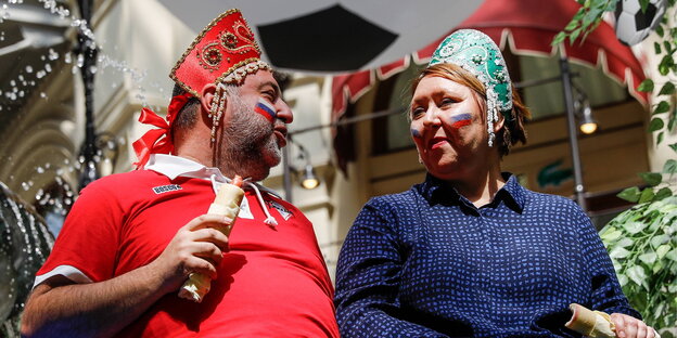 eine Frau und ein Mann mit traditionellem Kopfschmuck, die russische Fahnen auf die Backen geschminkt haben