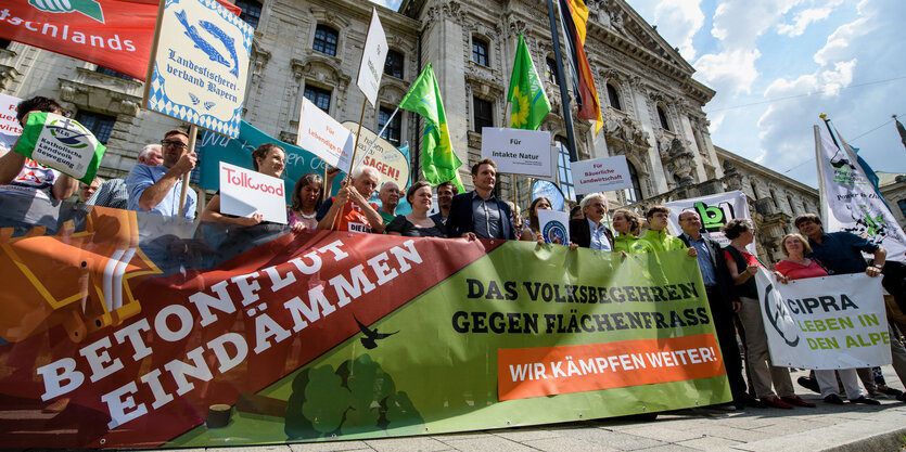 Demonstranten halten Transparente hoch, auf denen steht: Betonflut eindämmen. Das Volksbegehren gegen Flächenfraß