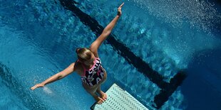 Eine junge Frau steht rückwärts auf einem Sprungturm, unter ihr ein Schwimmbecken