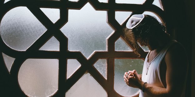 Ein Mann mit Kippa steht betend und mit gesenktem Kopf vor einem Fenster, dessen Streben einen Davidstern bilden