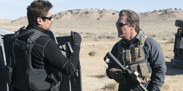 Benicio del Toro und Josh Brolin stehen in der Wüste und tragen schusssichere Westen und Waffen
