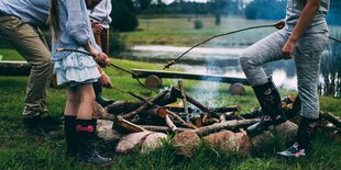 Mehrere Personen stehen an einem Lagerfeuer und grillen Lebensmittel auf Stöcken