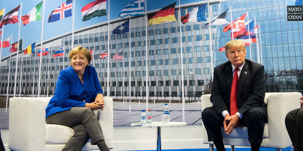 Angela Merkel und Donald Trump sitzen nebeneinander. Sie lächelt, er blickt grimmig