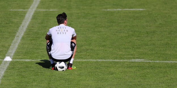 Mesut Özl sitzt auf einem Fußballplatz auf einem Ball, er ist von hinten zu sehen