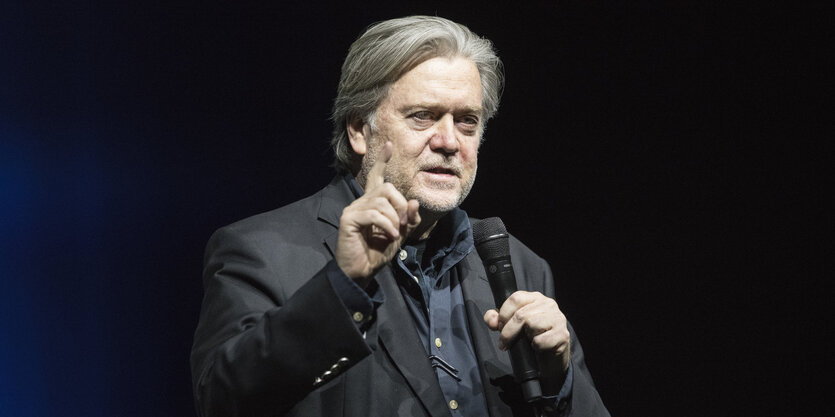 Steve Bannon hält ein Mikro in der einen Hand und streckt die andere mahnend mit erhobenem Zeigefinger hoch