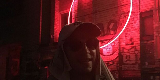 Ein Mensch mit Sonnenbrille in einem dunklen Raum, hinten an der Wand hängt eine rote Neonröhre in Kreisform