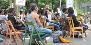 Viele Menschen sitzen auf Klappstühlen auf einer asphaltierten Straße, sie halten Schilder in die Luft. Auf einem steht: No Tranfer.