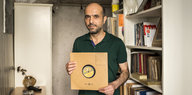 Ein Mann steht zwischen Regalen in seiner Wohnung und hält eine Schallplatte in altem braunen Papier in den Händen