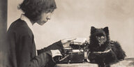 Eine junge Frau sitzt an einer Schreibmaschine, davor sitzt ein schwarzer Hund auf dem Tisch