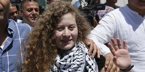 Junge Frau mit langen dunkelblauen Locken mit halberhobener Hand und Palästinensertuch um den Kopf. Im Hintergrund eine Menschenmenge
