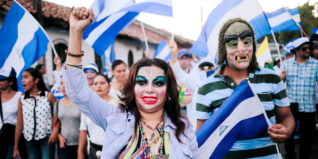 Eine übertrieben stark geschminkte Frau und ein mit einer Horrorfratze bemalter Mann marschieren bei einer Demonstration mit nicaraguanischen Fahrnen