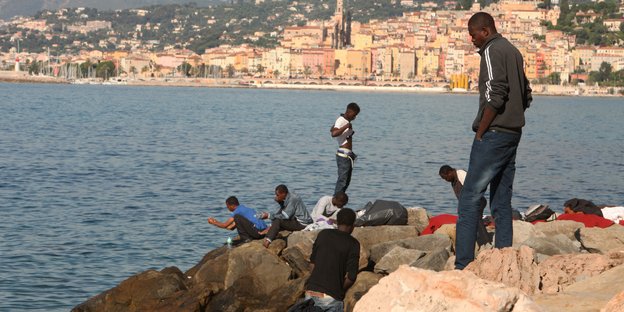 Menschen sitzen und stehen auf Steinen am Meer, im Hintergrund sieht man die italienische Stadt Ventimiglia