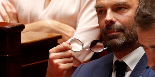 Edouard Philippe sitzt zwischen anderen Parlamentariern. Er hat sich die Brille abgesetzt und hält diese vor seinem Gesicht