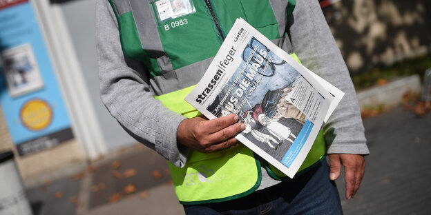 Zeitung Straßenfeger, gehalten von einer Person mit Warnweste hält