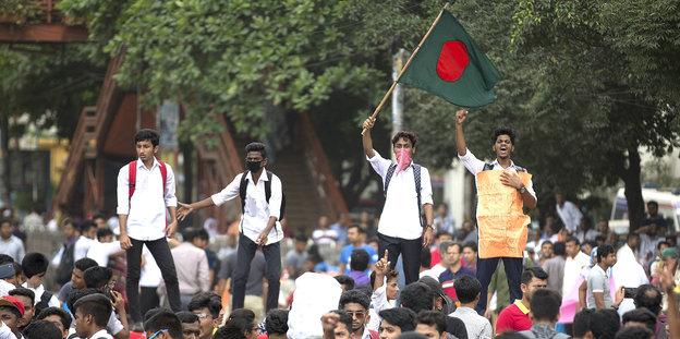 Vier junge Männer stehen erhöht über einer Menschenmenge, einer von ihnen schwenkt eine Fahne, zwei haben Tücher vor dem Mund