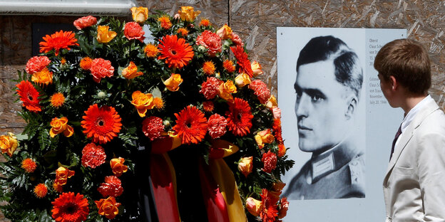 Eine Person steht vor einem Porträt von Graf Stauffenberg, sie ist nur von hinten zu sehen, neben dem Porträt sind Blumen drapiert