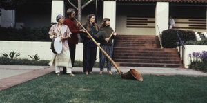 ein Mann mit einer Art Albhorn, eine Frau mit einer Trommel und zwei weitere Frauen stehen auf einer Wiese vor einem Haus