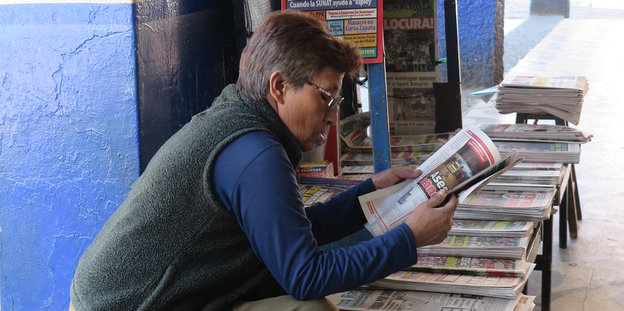 Frau sitzt über eine Zeitung gebeugt, lesend, dahinter ein Stand mit Zeitungen auf Spanisch