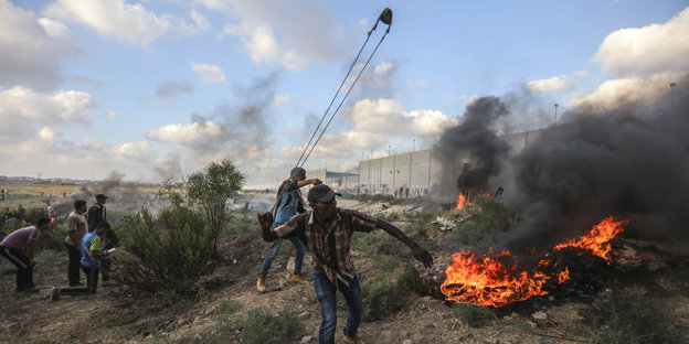An dem Grenzzaun zu Israel schleudert ein Mann einen Stein, daneben brennende Reifen