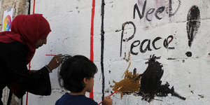 Eine Frau und ein kleiner Junge malen etwas an eine Wand. Zu lesen ist schon: "Need Peace"